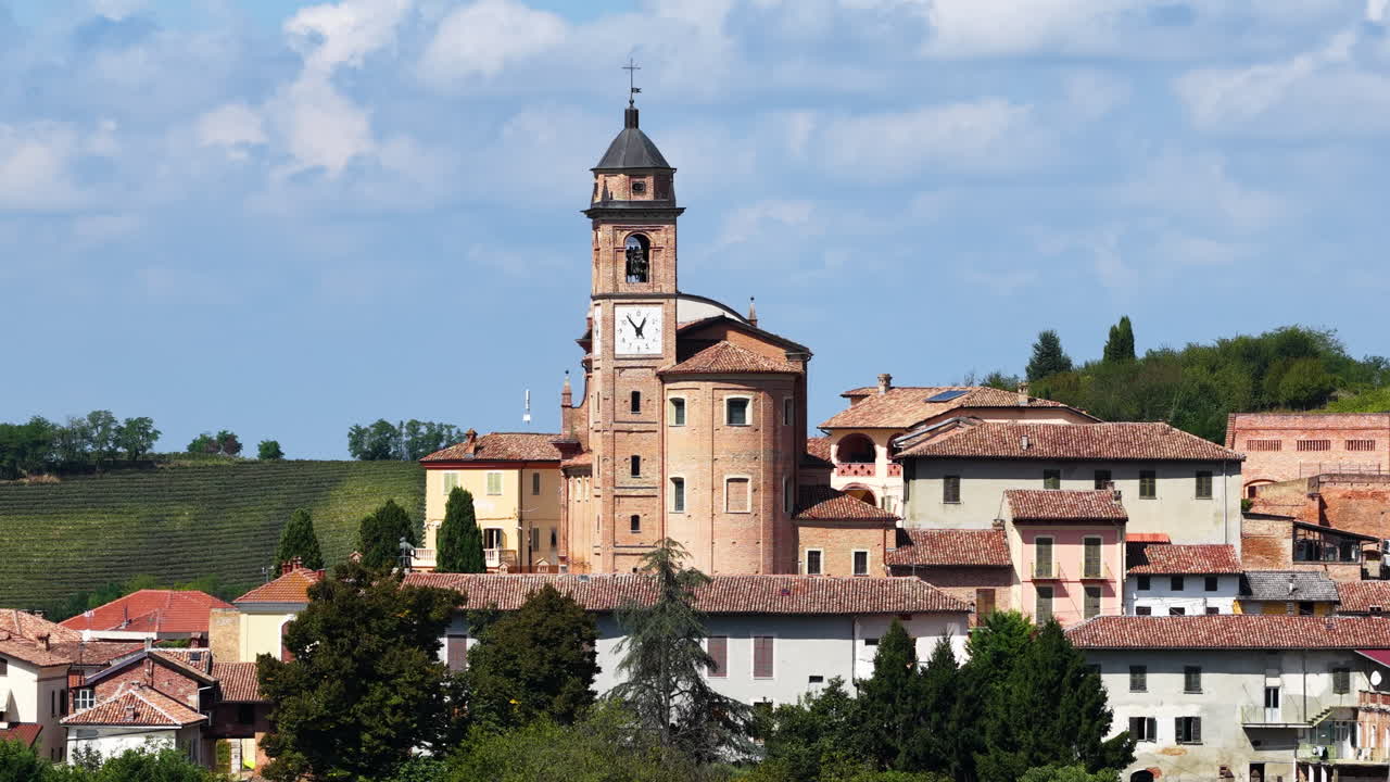 Ripresa cinematografica aerea con drone in parallax che rivela una chiesa storica circondata da case di paese e verdi colline nella regione Piemonte, nell'Italia settentrionale. Paesaggio europeo suggestivo