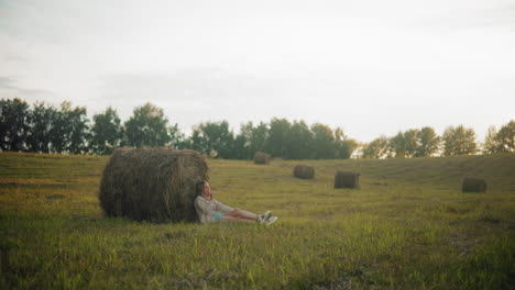 woman seated on ground thoughtfully, resting back against large hay bale in open farmland, peaceful rural landscape with scattered hay bales, warm evening light, and serene countryside setting