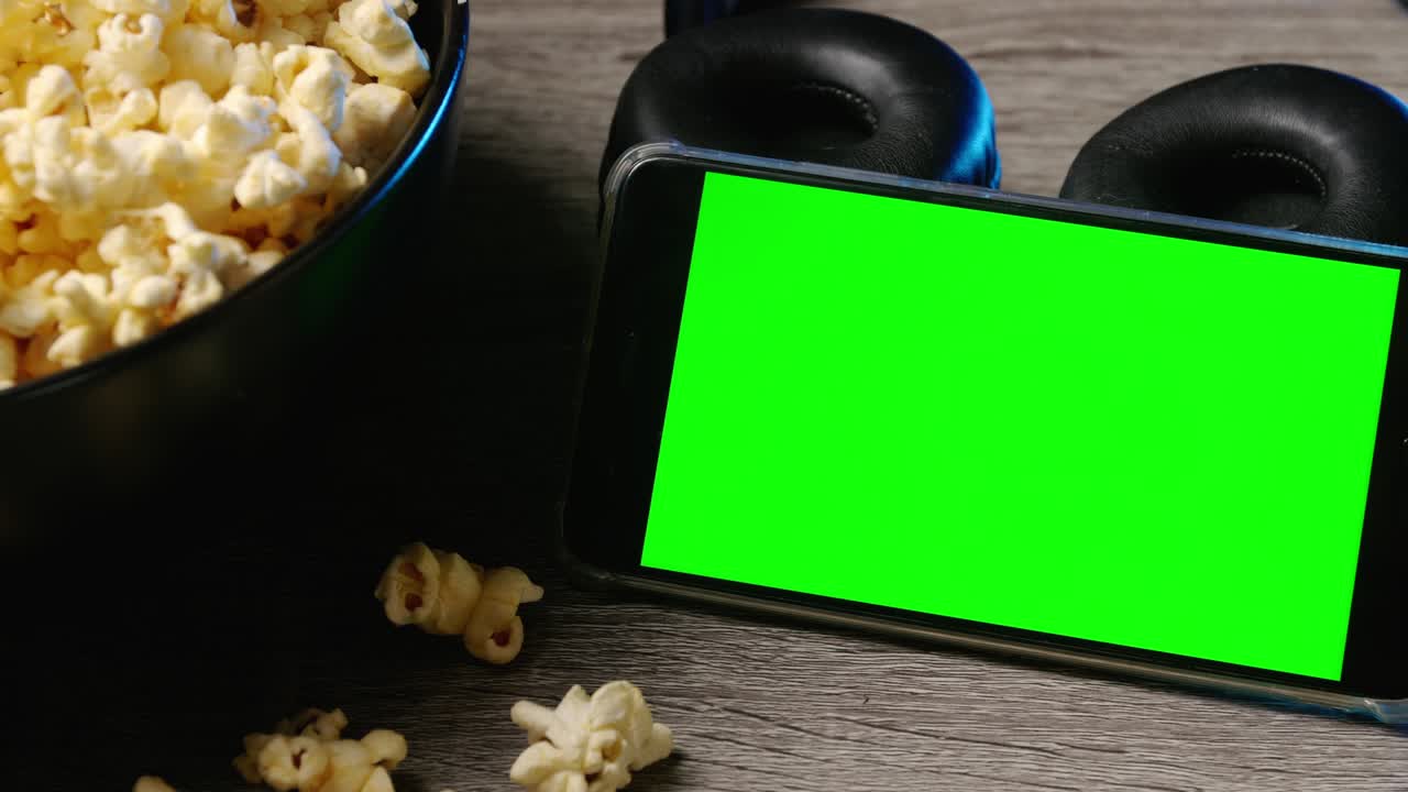 Smart phone with green touch screen and popcorn in a big bowl on the table.