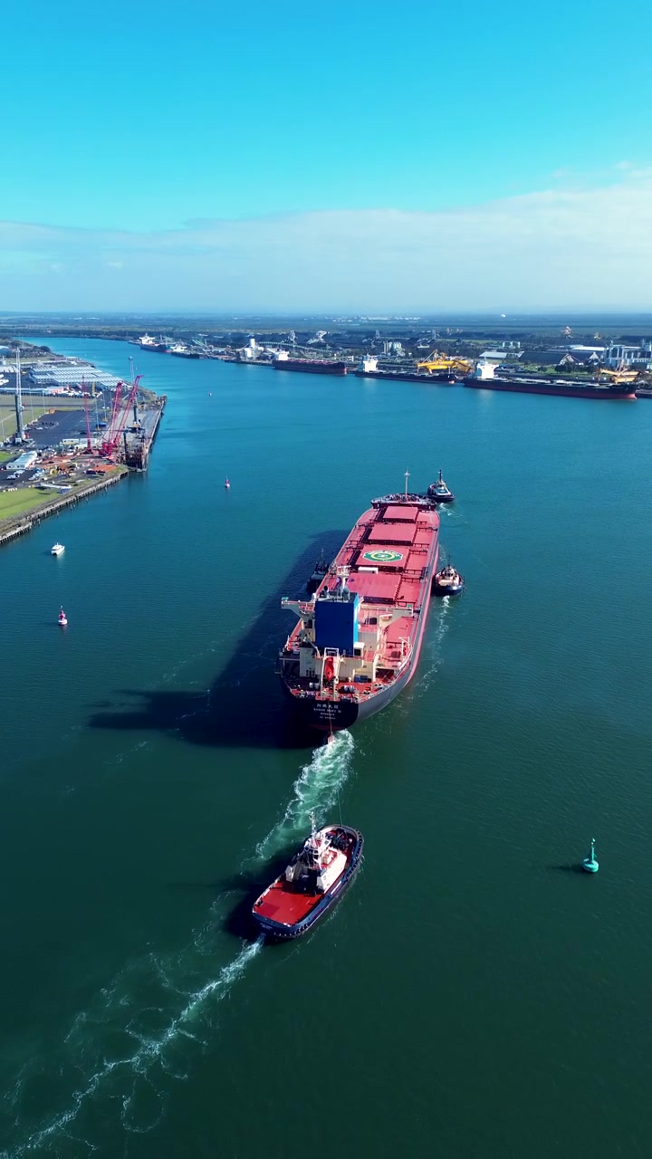 Retrato aéreo con dron de remolcadores guiando un buque de carga a granel a través del canal de agua del río Hunter en el puerto de la ciudad de Newcastle, con barcos amarrados en el entorno marítimo e industrial de Australia