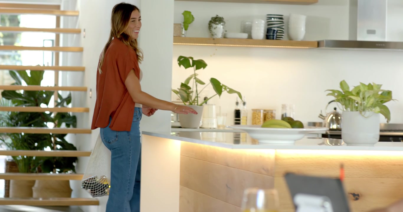 Famiglia che entra da sinistra con una donna che porta una borsa a rete, che disfa i bagagli sul bancone della cucina mentre un bambino guarda il tablet.