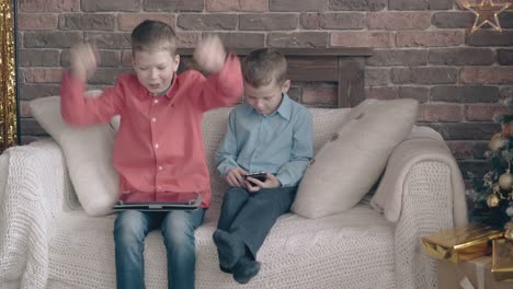 brothers sit on sofa playing on tablet and smartphone