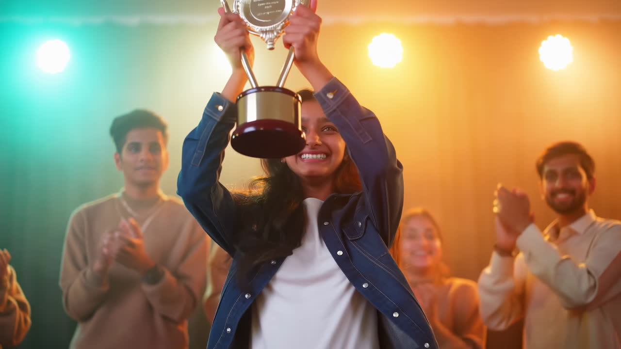 Donna indiana tiene un trofeo sul palco celebrando una vittoria gioiosa, studente sorridente solleva un premio sotto un palco colorato.