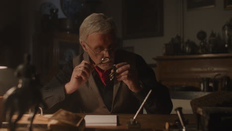 Older passionate man evaluates an old silver skull watch in his office at night