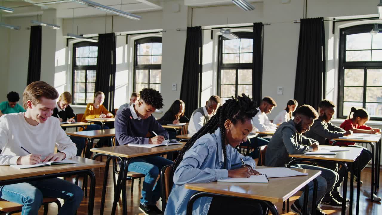High-angle video of diverse students focused on writing in a sunlit classroom, capturing a studious