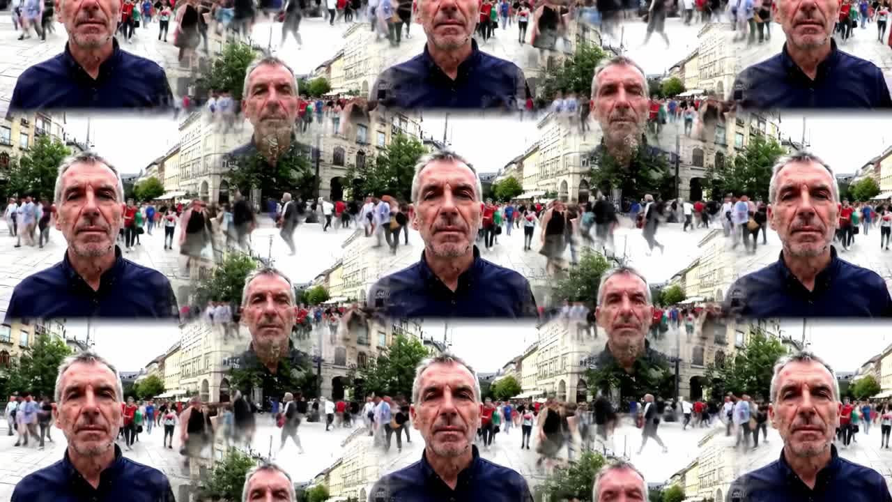 A man stands still in a bustling city plaza as people walk by in various directions. The surrounding architecture creates a vibrant atmosphere filled with movement and energy.