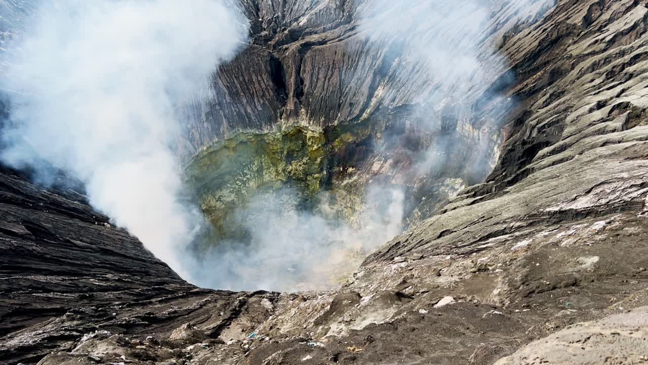 Dramatic view into the active Mount Bromo crater in East Java Indonesia, reveals thick volcanic smoke rising from steep mineral stained walls formed by powerful eruptions and ongoing volcanic activity