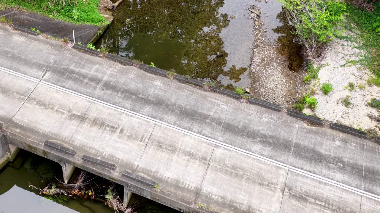 A white car drives across a concrete bridge over a river, surrounded by dense greenery, captured from above