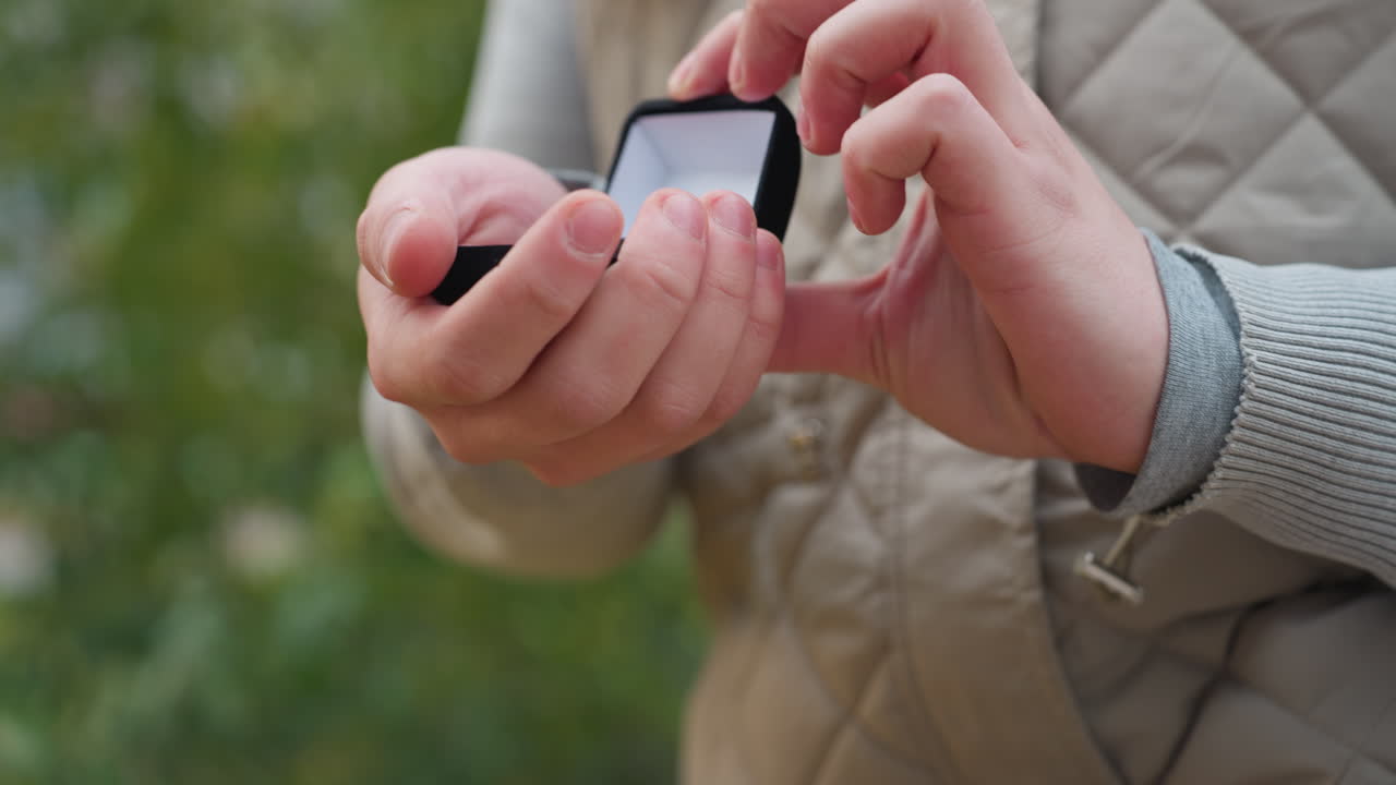 Close up of young man in quilted jacket preparing for proposal on one knee while holding ring gently in hands with soft focus background of greenery and subtle autumn tones