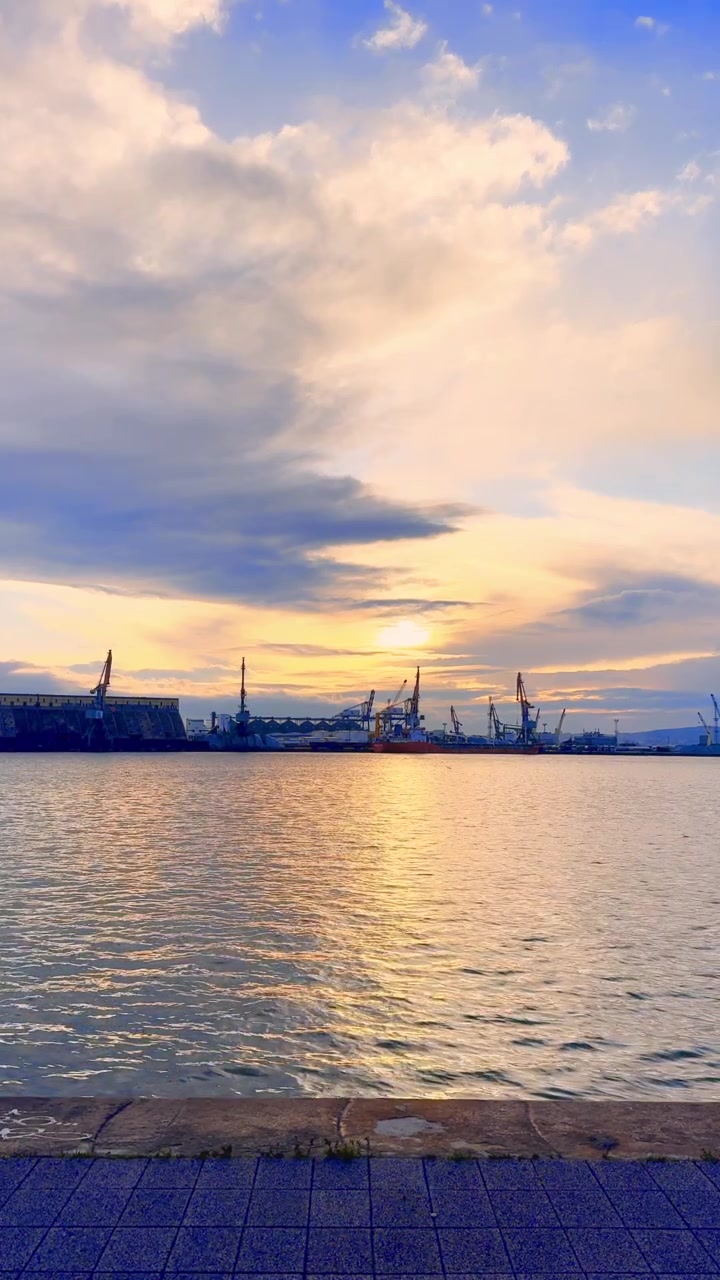 Sunset view over the harbor with boats. The sun sets behind the harbor, casting colorful light on the water. Boats and cranes are visible in the distance.