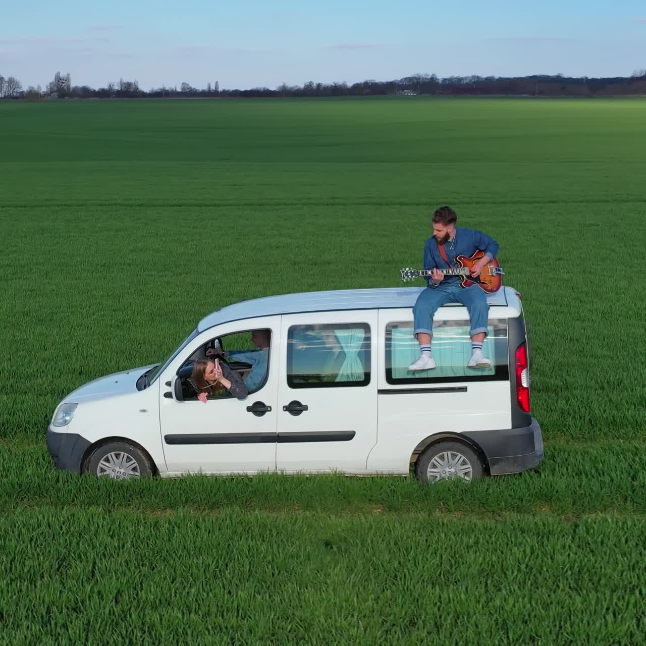 Young man playing the guitar on a moving car. Friends in white car travel on green field while guitarist sitting on a roof of the auto. Extreme for young people