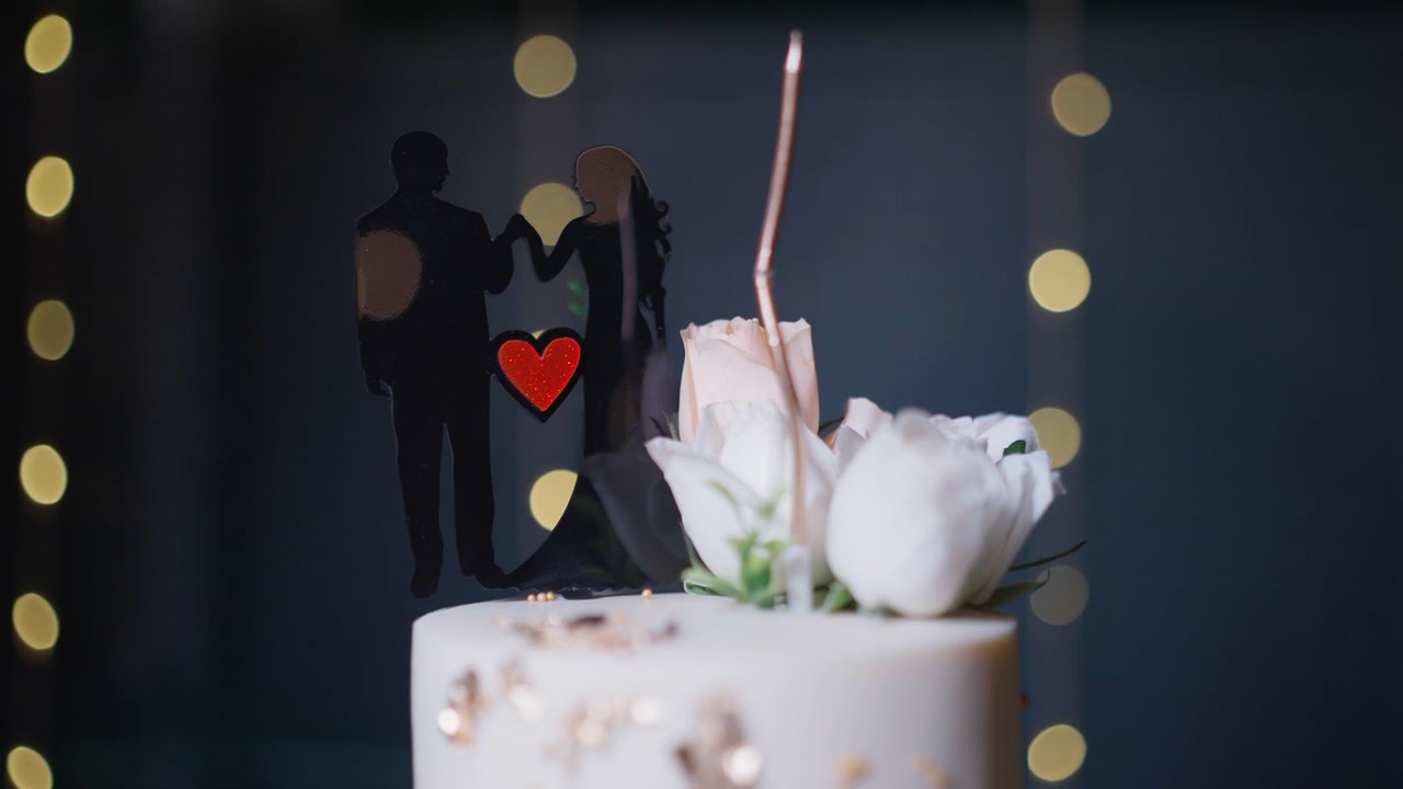 Primo piano di un cake topper nuziale che mostra la silhouette di una coppia con cuore rosso e fiori bianchi contro luci bokeh soffuse a una celebrazione pre-matrimoniale Sangeet