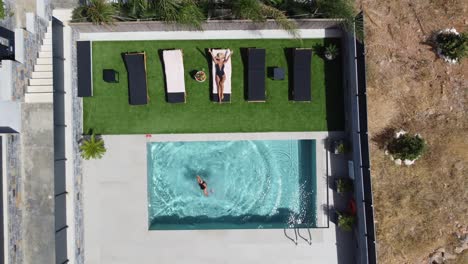 drone flies over an outdoor pool in bird's eye view - woman in swimming costume lies on sunbed and man jumps into the pool - villa in greece crete with palm trees