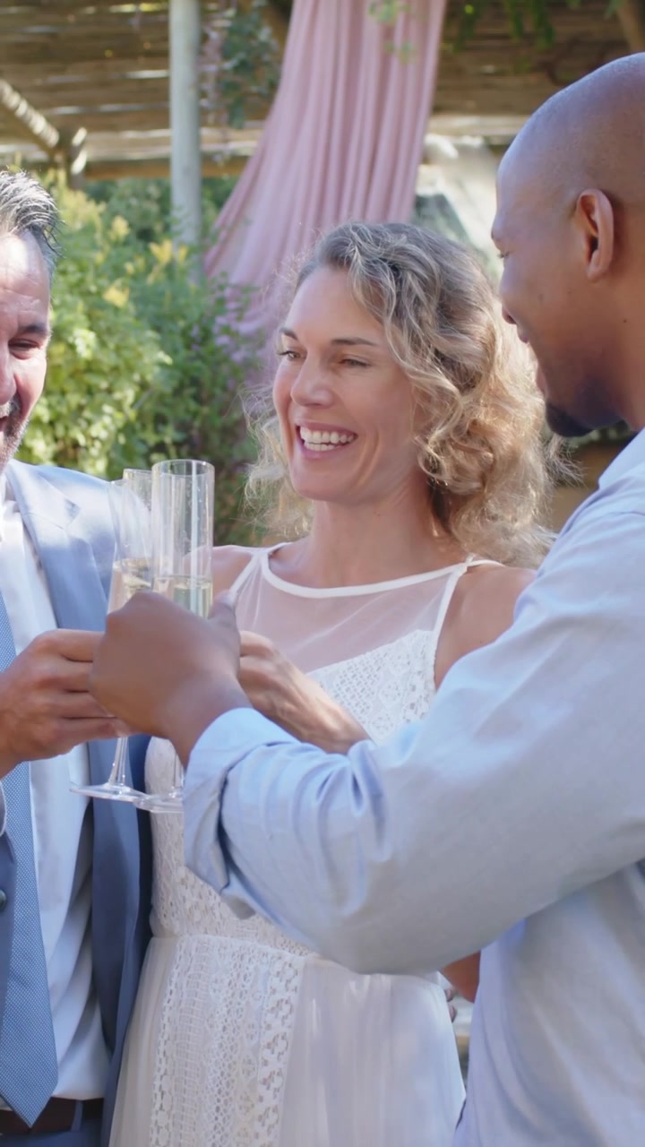 Vídeo vertical: Levantando taças, noiva começando brinde de casamento no jardim com homens de terno e camisa