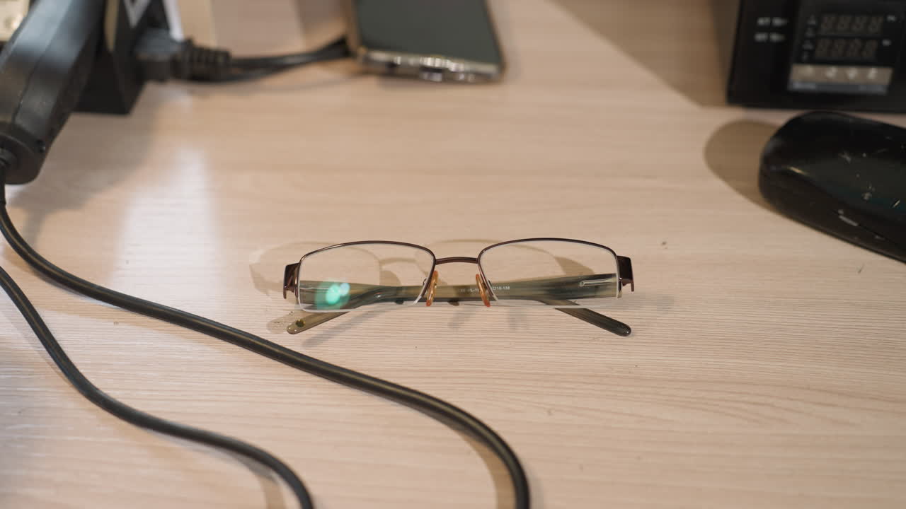 A close-up view of someone right hand wearing a black glove reaching for a pair of glasses on a wooden desk, with phone on the table and other equipment