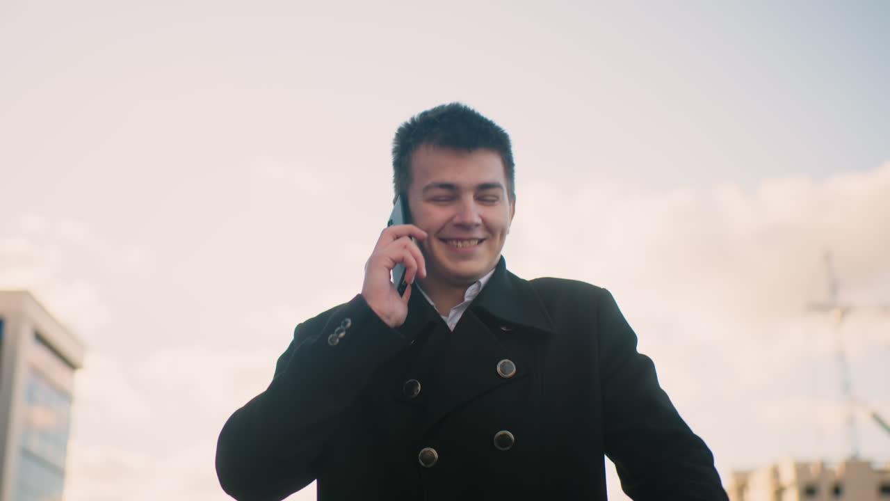 현대적인 유리 건물, 부드러운 빛, 그리고 즐거운 분위기가 있는 활기찬 도시 지역에서 웃고 흥분한 손짓을 하면서 전화로 이야기하는 검은 코트를 입은 남자