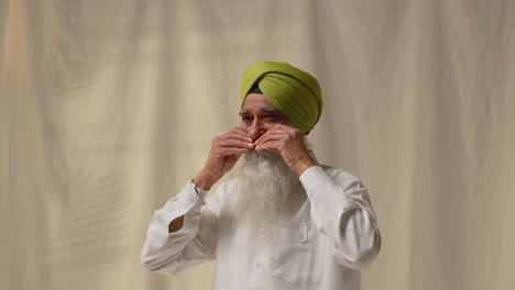 studio shot of senior sikh man grooming beard and moustache checking turban against plain background