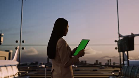 vista trasera de una mujer de negocios asiática con una maleta rodante en la sala de embarque del aeropuerto, usando una tableta de pantalla verde mientras espera el vuelo, el avión despega fuera de la ventana