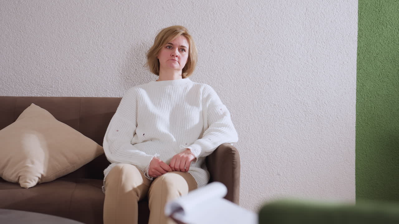 Patient seated on sofa opposite professional nodding approval during therapy session in modern cozy room with pillow, knit blanket, wooden table visible in neutral wall background soft lighting