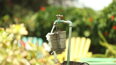 water from a garden spout pours and spills over a hanging bucket into a larger barrel with colored lounge chairs in the background