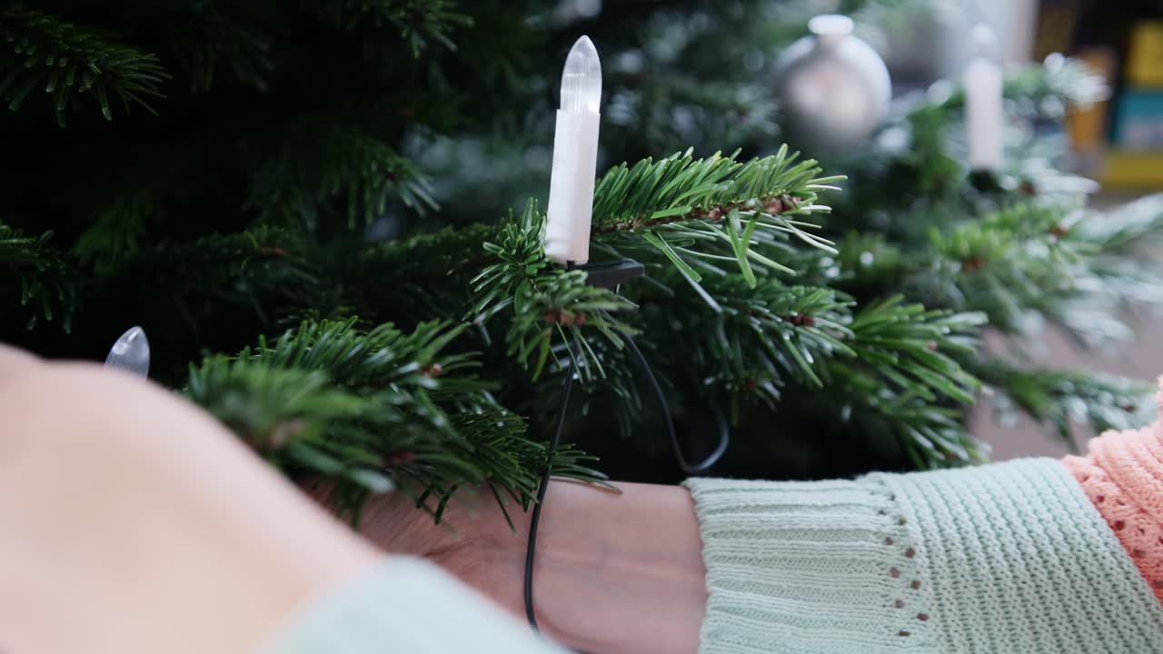 Over-the-shoulder close-up of Caucasian hands placing LED candles on a Christmas tree. Soft foreground blur adds depth, creating a calm, traditional decorating moment