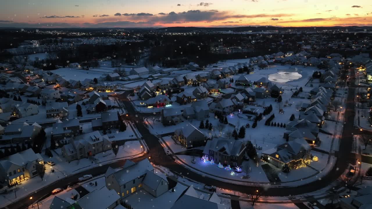 Laaja ilmakuva kodikkaasta talvimaisemasta, jossa lumipyry auringonlaskun aikaan. Hehkuva julkisivu sekä talojen ja kotien pihat jouluisena aikana. Rauhallinen naapurusto. Hehkuvat lämpimät lyhtyvalot