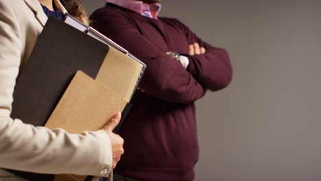 close up studio shot of male and female teachers standing against grey background holding folder under arm 1