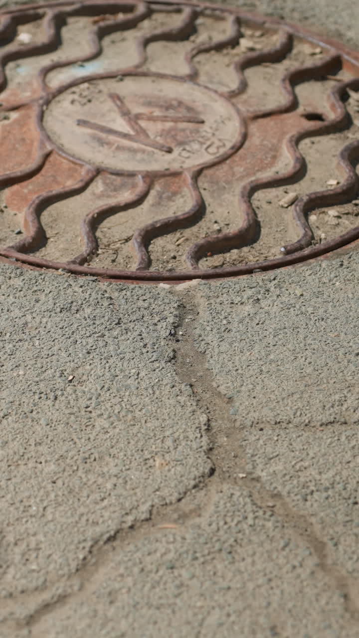 Close view of a manhole cover on a cracked asphalt road, partially obscured by a shadow, with people passing by and there shadows casted on the ground