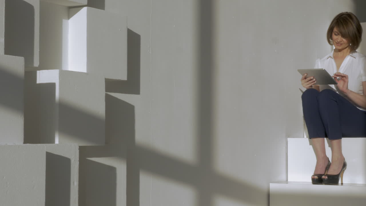 젊은 사업가 여성이 계단에 앉아 태블릿에 타이핑하고 미소 짓고 있습니다.