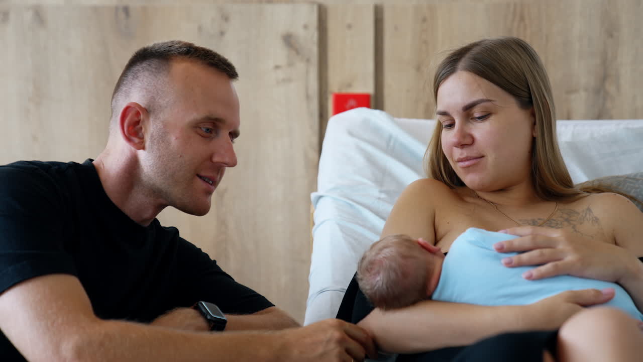New parents in the maternity hospital ward after labor. Man touching tenderly his newborn lying in mom's hands. Family portrait.