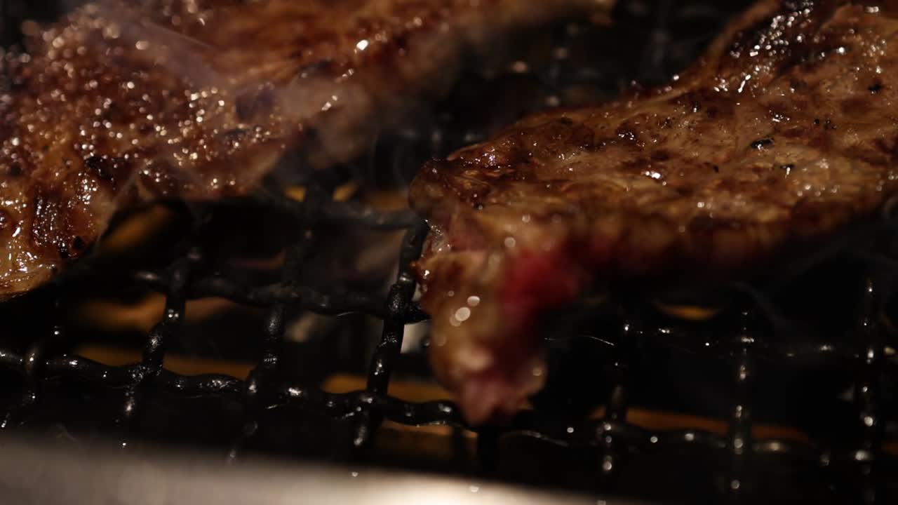 Close-up of a steak sizzling on a grill with smoke rising and flames visible beneath.