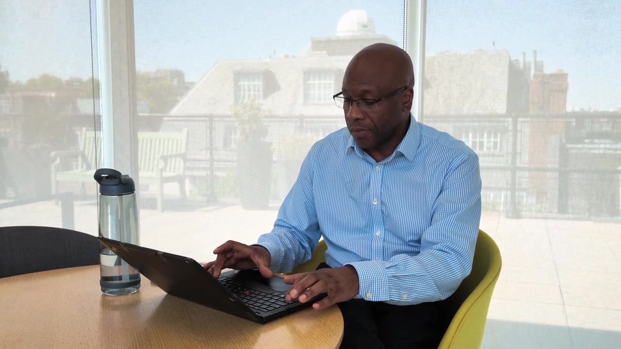 Corporate employee sits at a round table using his smartphone beside an open laptop and water bottle in a sunlit meeting room.