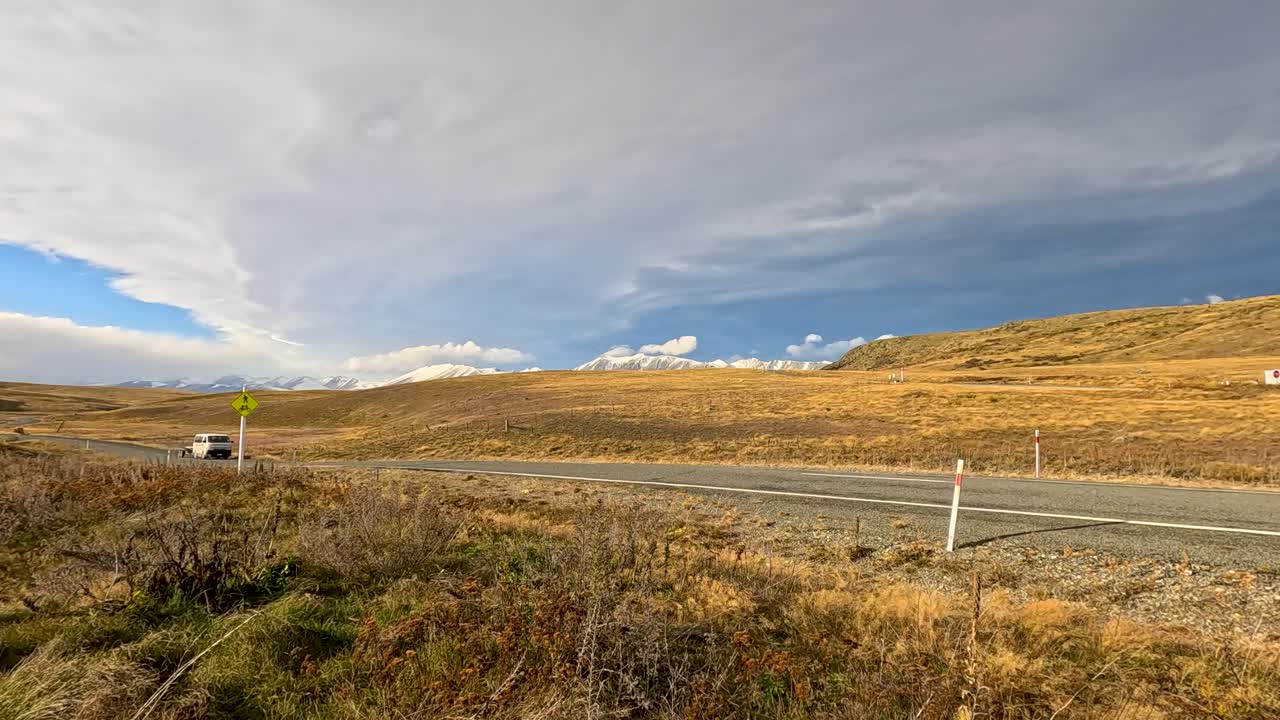 A white camper van travels on a rural highway through golden autumn grasslands under dramatic skies near Lake Tekapo, New Zealand. Static wide shot, natural daylight