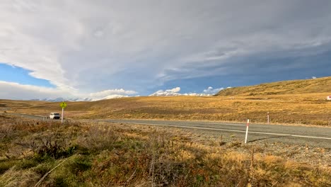 a white camper van travels on a rural highway through golden autumn grasslands under dramatic skies near lake tekapo, new zealand. static wide shot, natural daylight
