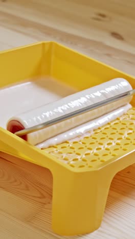 vertical close-up of a paint roller being evenly coated with white paint in a bright yellow tray, placed on a wooden floor, showing the preparation phase of an indoor renovation project