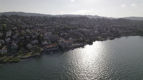 Drone-shot-flying-near-the-city-of-Thalwil-at-the-Zurich-Lake-in-Switzerland-on-a-sunny-day-with-water-and-boats-around-the-shore-and-white-houses-in-the-city-LOG