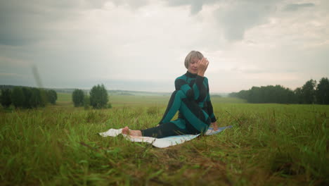 advanced woman in green black suit seated on yoga mat practicing half seated twist, gently lifting head backwards, in a vast grassy field with trees in the distance under cloudy sky