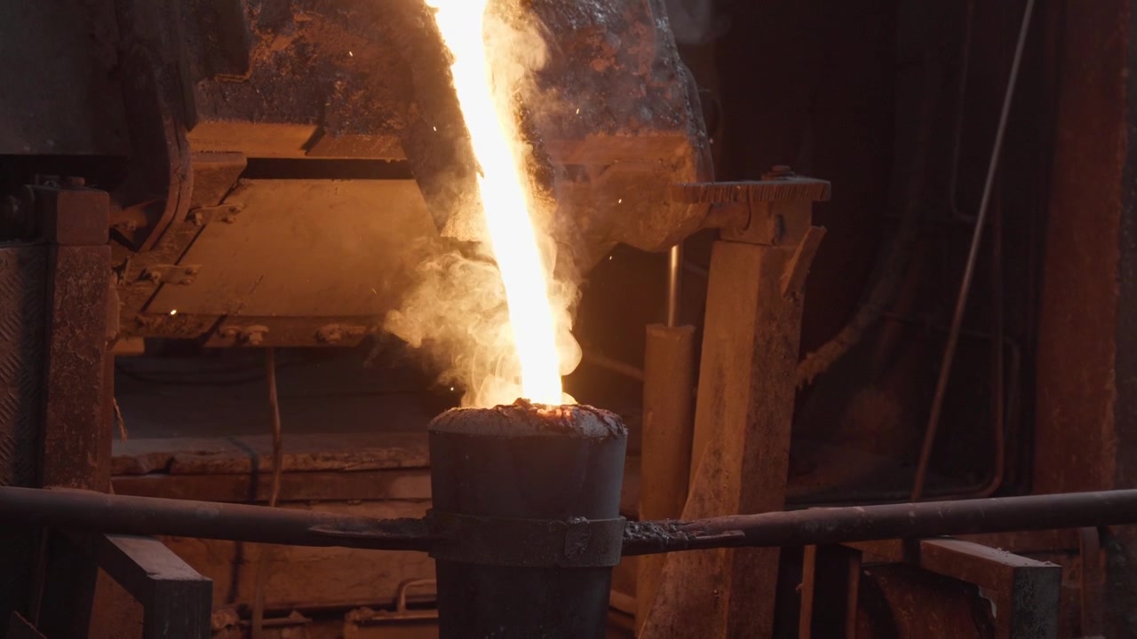 Un flusso luminoso inizia a scorrere dal beccuccio della fornace in un mestolo a manico lungo, con fumo che si alza dal recipiente di raccolta