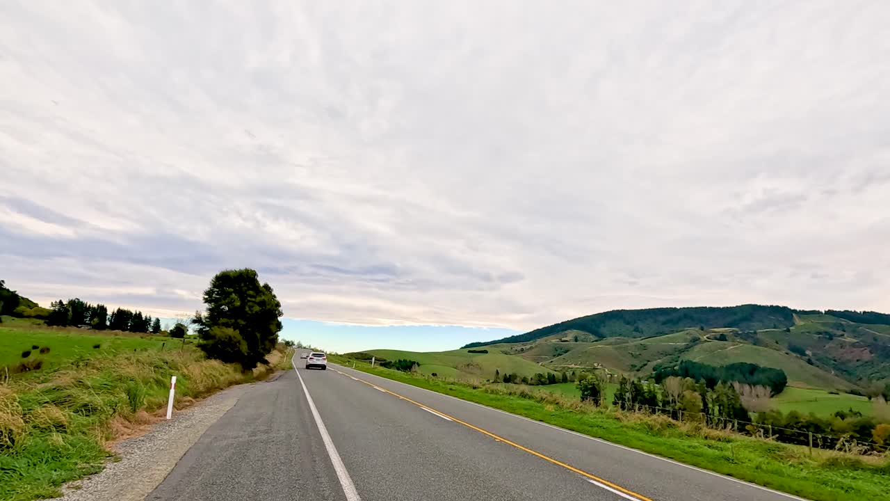 A car travels along a rural road with rolling hills and cloudy skies at Lake Tekapo, New Zealand