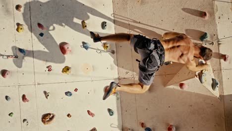 Vertical-video-of-a-brunette-guy-in-Gray-shorts-with-a-bare,-sculpted-torso-climbs-up-the-route-of-a-climbing-wall-and-hangs-on-one-of-the-ledges-with-the-help-of-a-belay-and-his-strong-arms-during-a-rock-climbing-training-at-a-climbing-wall-in-the-summer