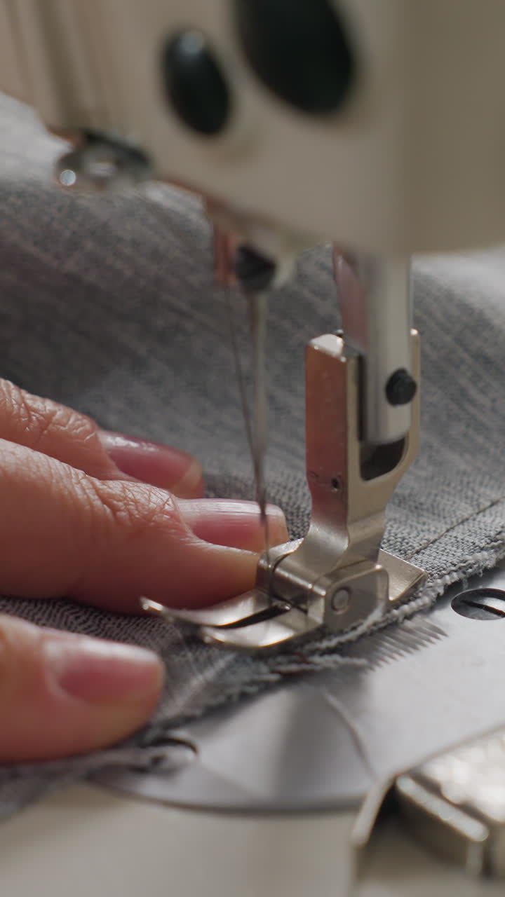 Primer plano de una costurera asiática cosiendo tela vaquera en una máquina industrial, con los dedos guiando la tela gris bajo el prensatelas, la aguja perforando la tela para crear puntadas precisas, detalle de botón visible, taller cálido