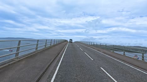 a vehicle travels along a two-lane bridge with metal railings, surrounded by water and distant hills under bright, overcast daylight. smooth forward camera movement