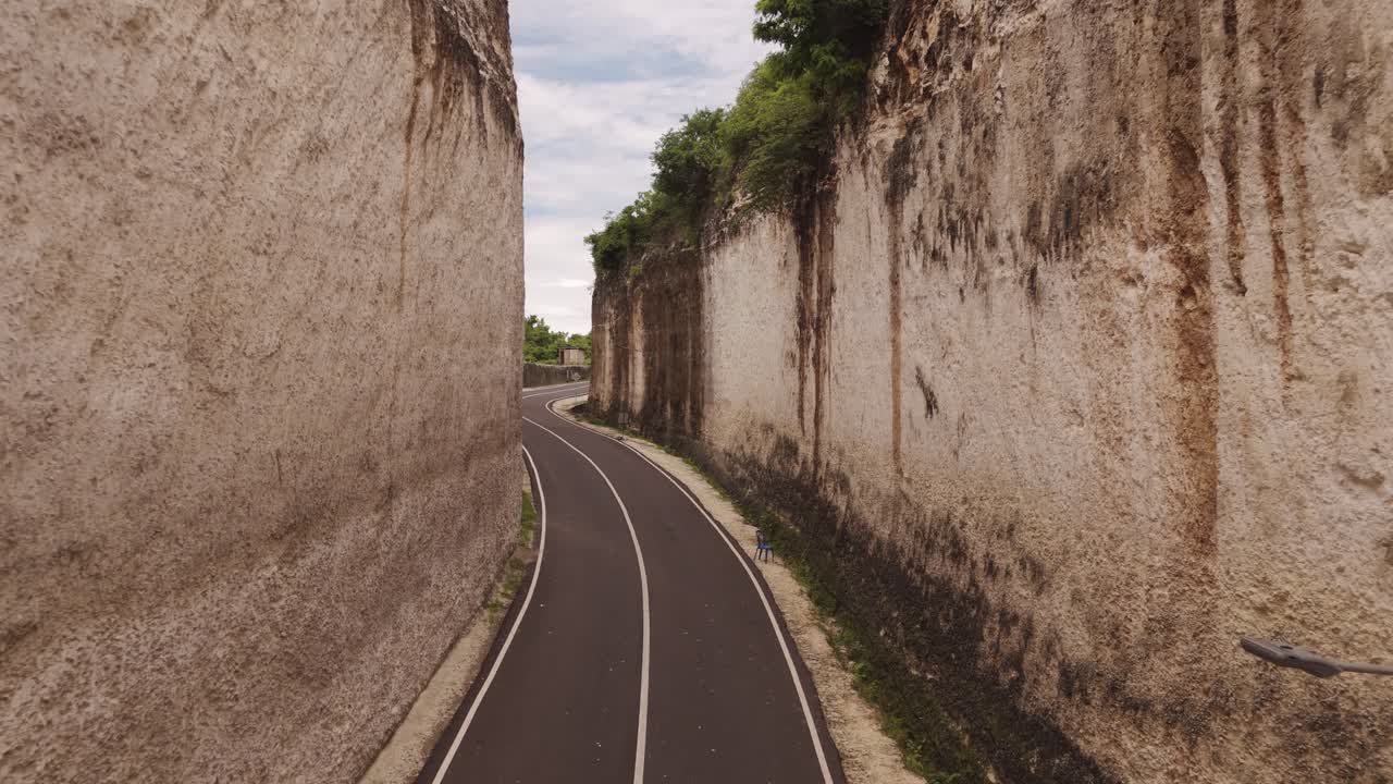 Cinematic low aerial shot gliding above a narrow, empty road between towering stone cliffs at Tanah Barak, Bali, Indonesia—showcasing stunning natural formations and peaceful tropical scenery.