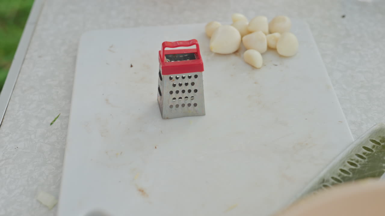 Aglio preparato e disposto all'aperto. Ambiente esterno sereno con spicchi d'aglio e grattugia sull'erba. Tranquilla scena di picnic che mostra aglio e utensili da cucina in un ambiente esterno erboso