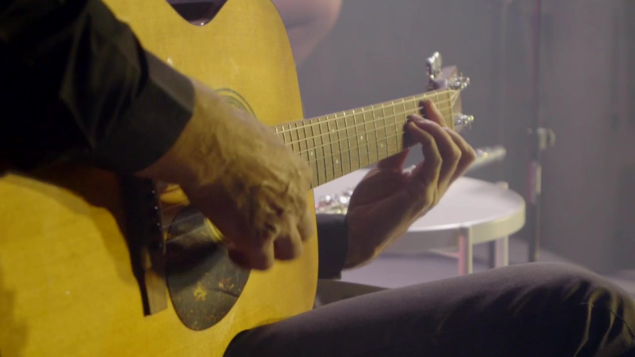 En mand spiller akustisk guitar på scenen, med et nærbillede af hans hænder og guitarstrengene.