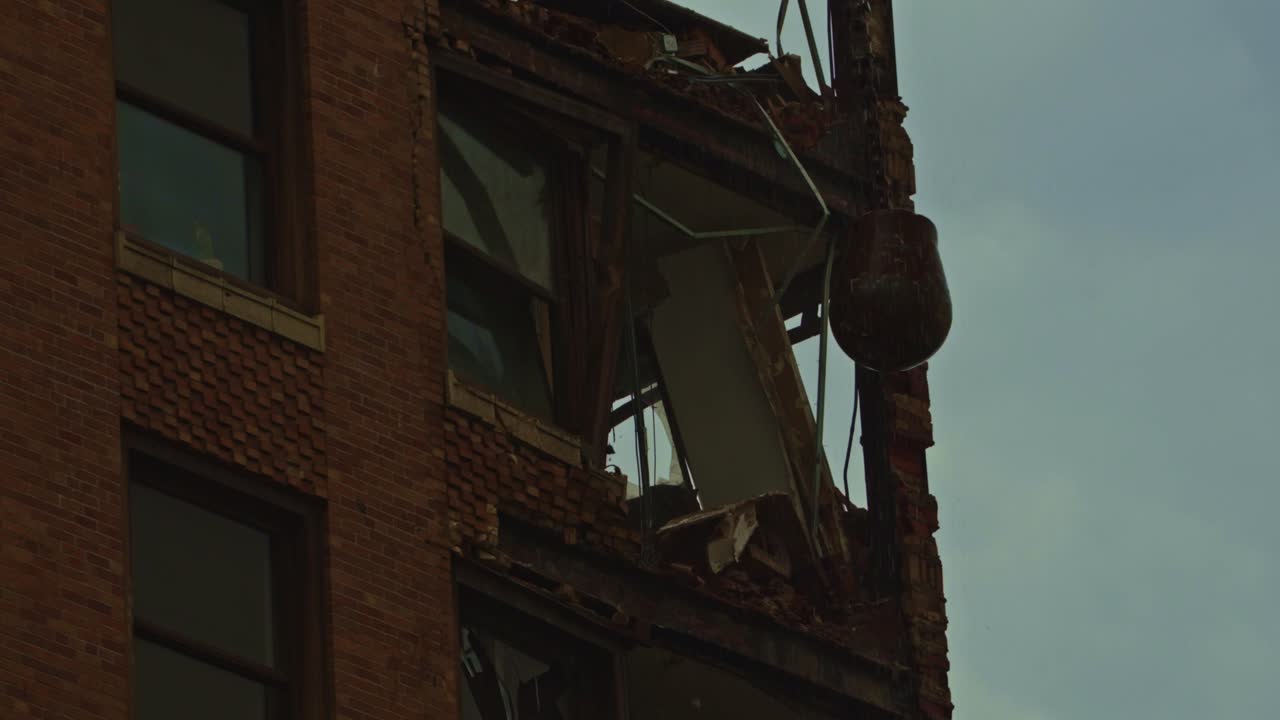 Zoomed-in view of a wrecking ball smashing into the side of a red brick high-rise, shattering windows and interior walls during demolition.