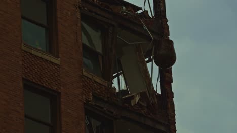 zoomed-in view of a wrecking ball smashing into the side of a red brick high-rise, shattering windows and interior walls during demolition.