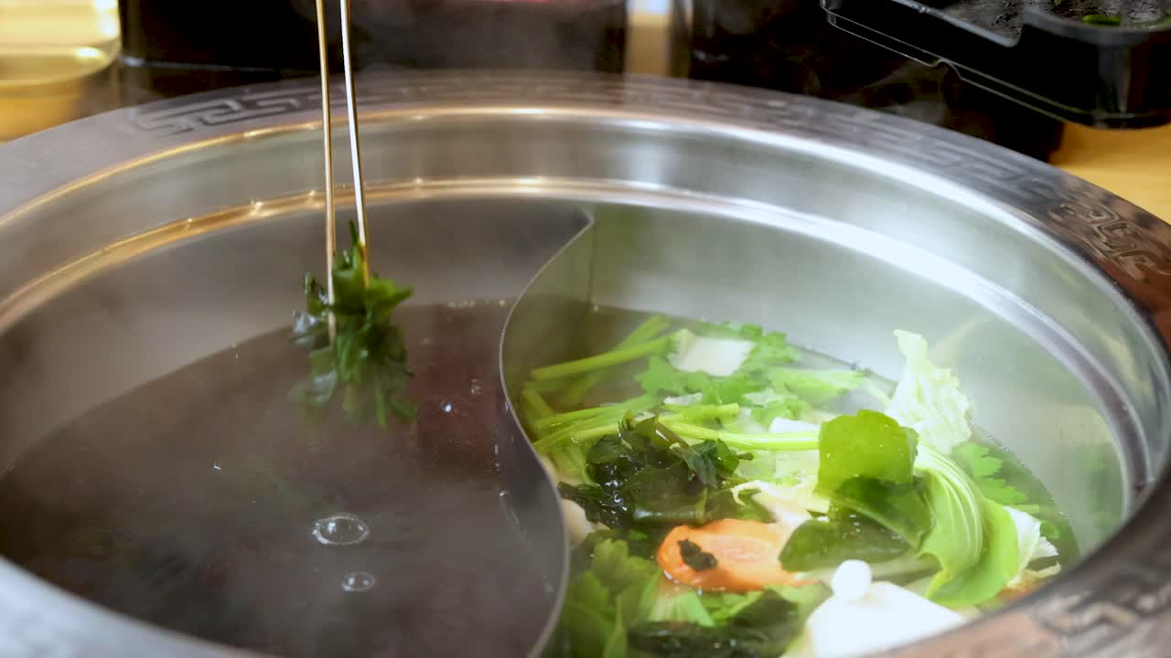 Chopsticks stir fresh vegetables in steaming dual broth hotpot, indoor dining, bright lighting, close-up