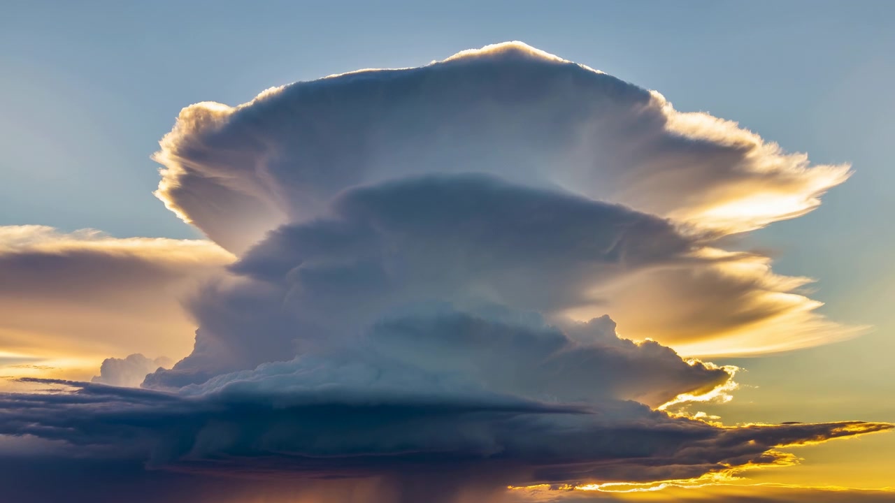 Massiv cumulonimbussky fylder den brede himmel med lagdelt amboltformet top, afrundede tårne, skarpt kantlys og synlige regnskyer over den mørke horisont under varmt solnedgangslys, hvilket skaber en kraftfuld atmosfære.