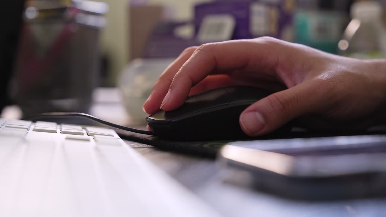 a person's hand using a black computer mouse on a desk with a white keyboard and smartphone. Shallow depth of field in a home office during the day